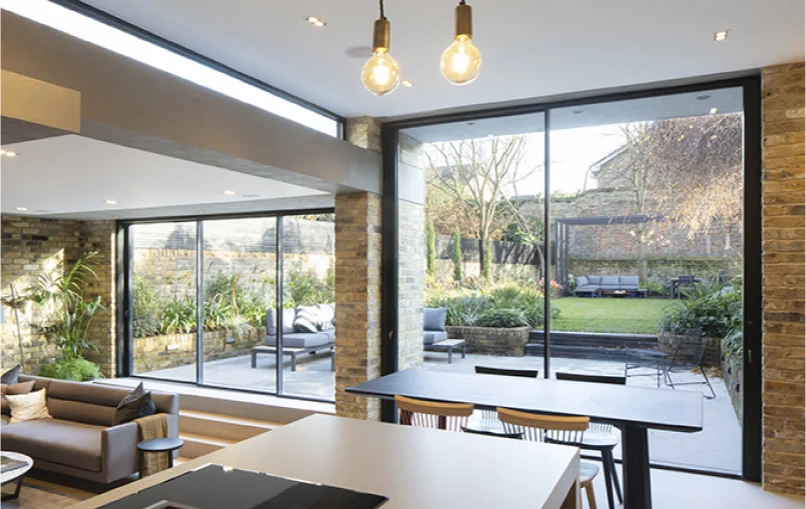 This image depicts the view from the kitchens island. From the island you can see the living space in the previous picture and a dinign table. Beyond the dining table are two verticle windows that block no sunlight from entering the house and allow perfect view of the garden.