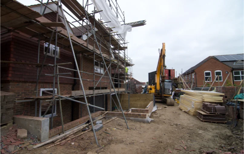 This image depicts the parking lot for the old office building which now has 2 large, 3 storey houses built in its place with a 15ton JCB digger parked outside surrounded by building material.