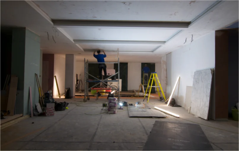 This image depicts the whole basement under construction with plaster boards on the walls and ceilings and an electrician mounting LED strips to the ceiling.