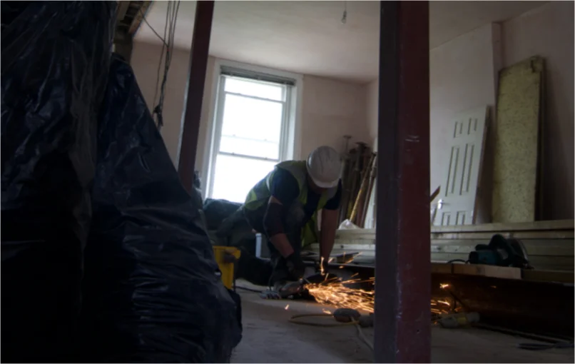 This image depicts a construction worker with a hi-vis jacket and a white helmet using an angle grinder to cut a steel beam.