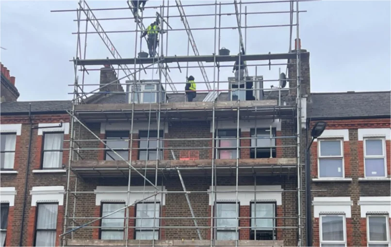 This image depicts the outside of an old 4 storey building in London with scaffold workers working on the top floor putting up the last pieces of scaffold.